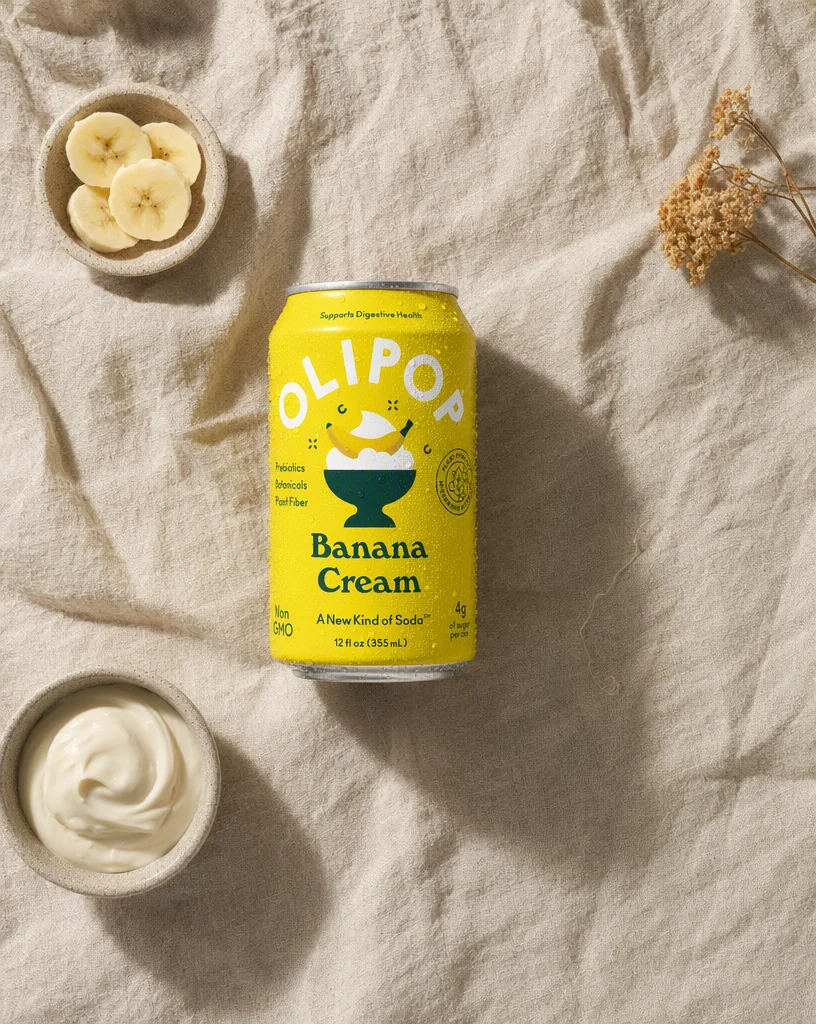 Overhead flat-lay of the OLIPOP can on linen with a dish of banana slices, cream and dried flowers
